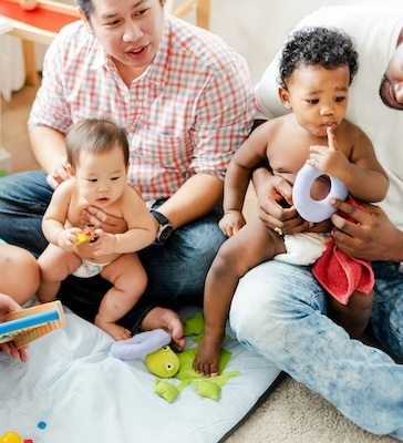 Two fathers playing on the floor with their babies and toys