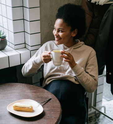 Smiling pregnant woman in a beige hoodie holding a mug of coffee at a café table