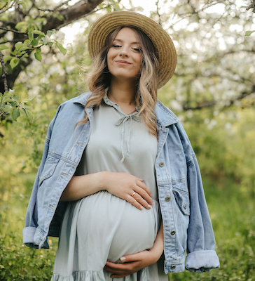 Smiling pregnant woman in a straw hat and denim jacket standing in a sunlit orchard