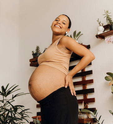 Smiling pregnant woman in a beige tank top resting one hand on her lower back